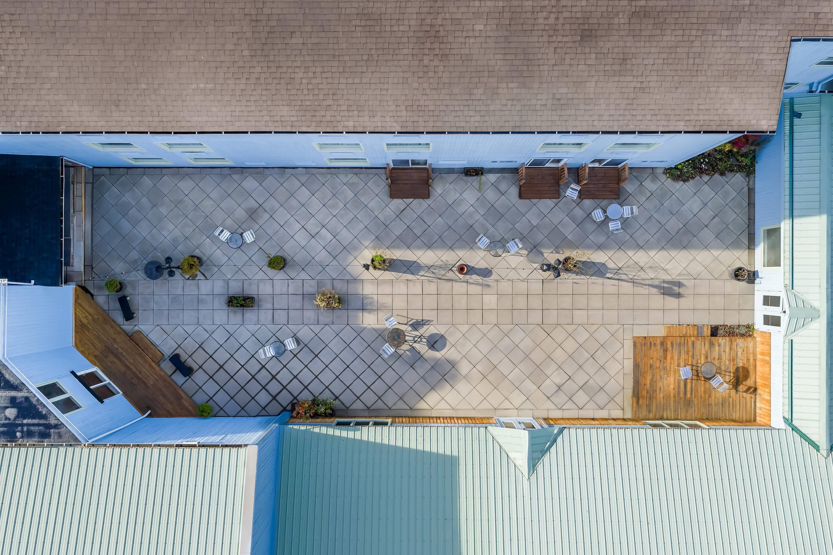 Vista aérea de un patio con varias zonas de estar, incluyendo mesas y sillas, rodeado de un suelo de baldosas y jardineras de madera.