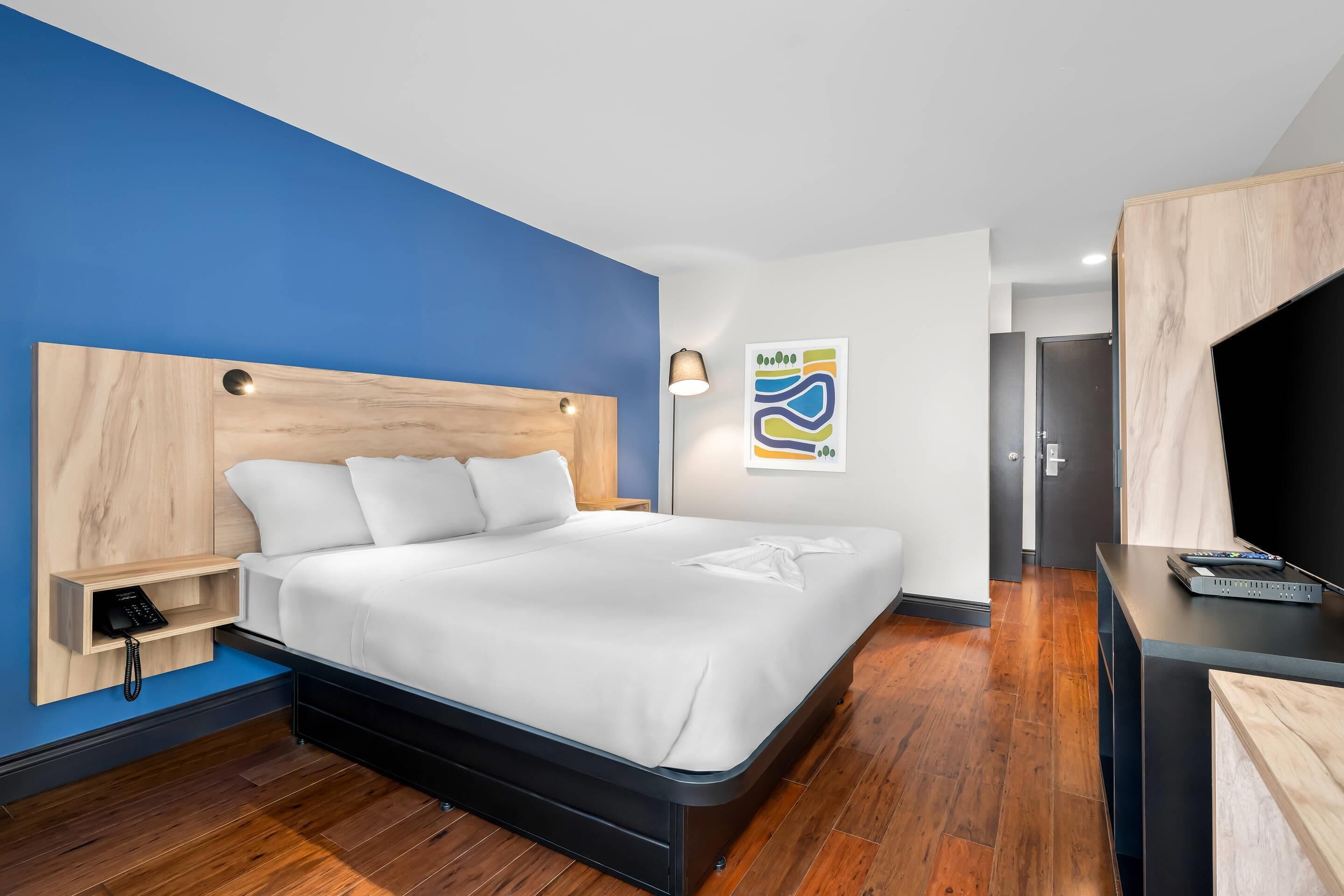 A modern hotel room with a large bed, a bedside table with a phone, a wall-mounted artwork, and a TV on a stand.