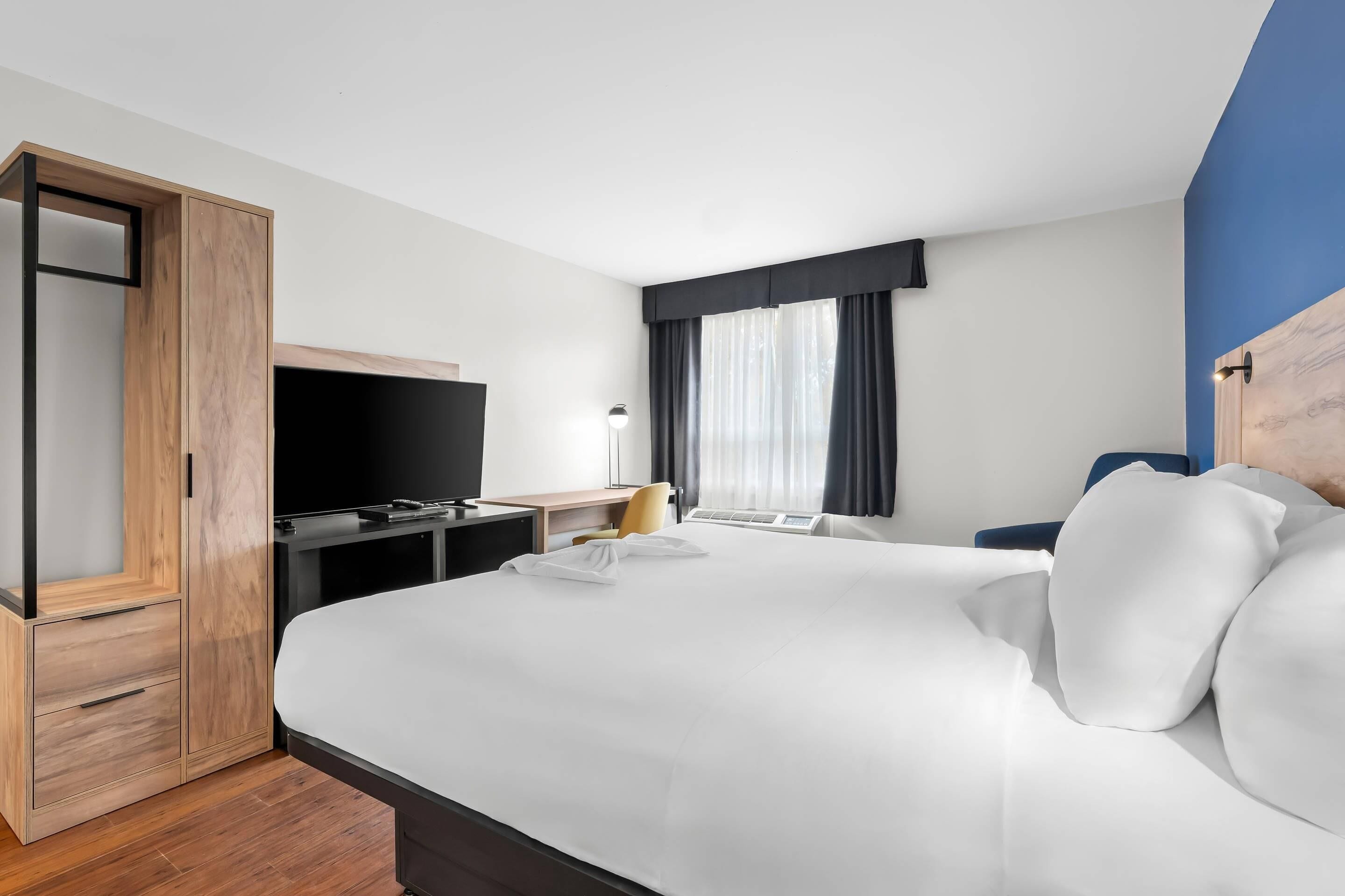 Une chambre d’hôtel avec un grand lit, un bureau doté d’un téléviseur à écran plat, une armoire et une fenêtre avec des rideaux.
