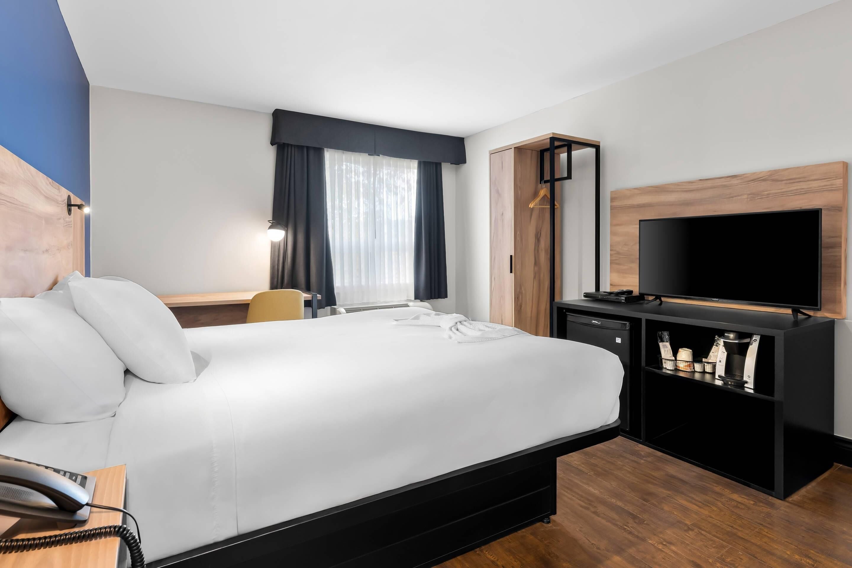 Habitación de hotel con una cama grande, un escritorio con teléfono, un televisor sobre un soporte de madera y una ventana con cortinas.