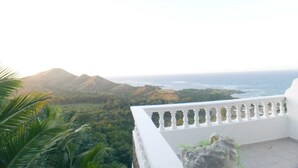 Un balcon doté d’une balustrade blanche, offrant une vue sur un paysage côtier avec des montagnes et l’océan.