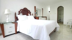 Une chambre à coucher bien rangée, avec une tête de lit en bois, une table de chevet en verre, une lampe blanche, un miroir et une petite chaise blanche.