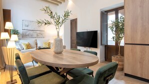 Ein modernes Wohnzimmer mit einem runden Holztisch, grünen Polsterstühlen, einem großen Flachbildfernseher und einer Zimmerpflanze.