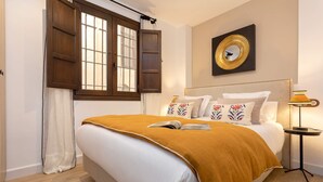 Una cama bien hecha con un edredón color mostaza, una mesita de noche con lámpara y un espejo en la pared.