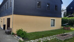 A two-story house with a dark roof and a light beige wall. There is a bench and a small garden area with a brick planter.