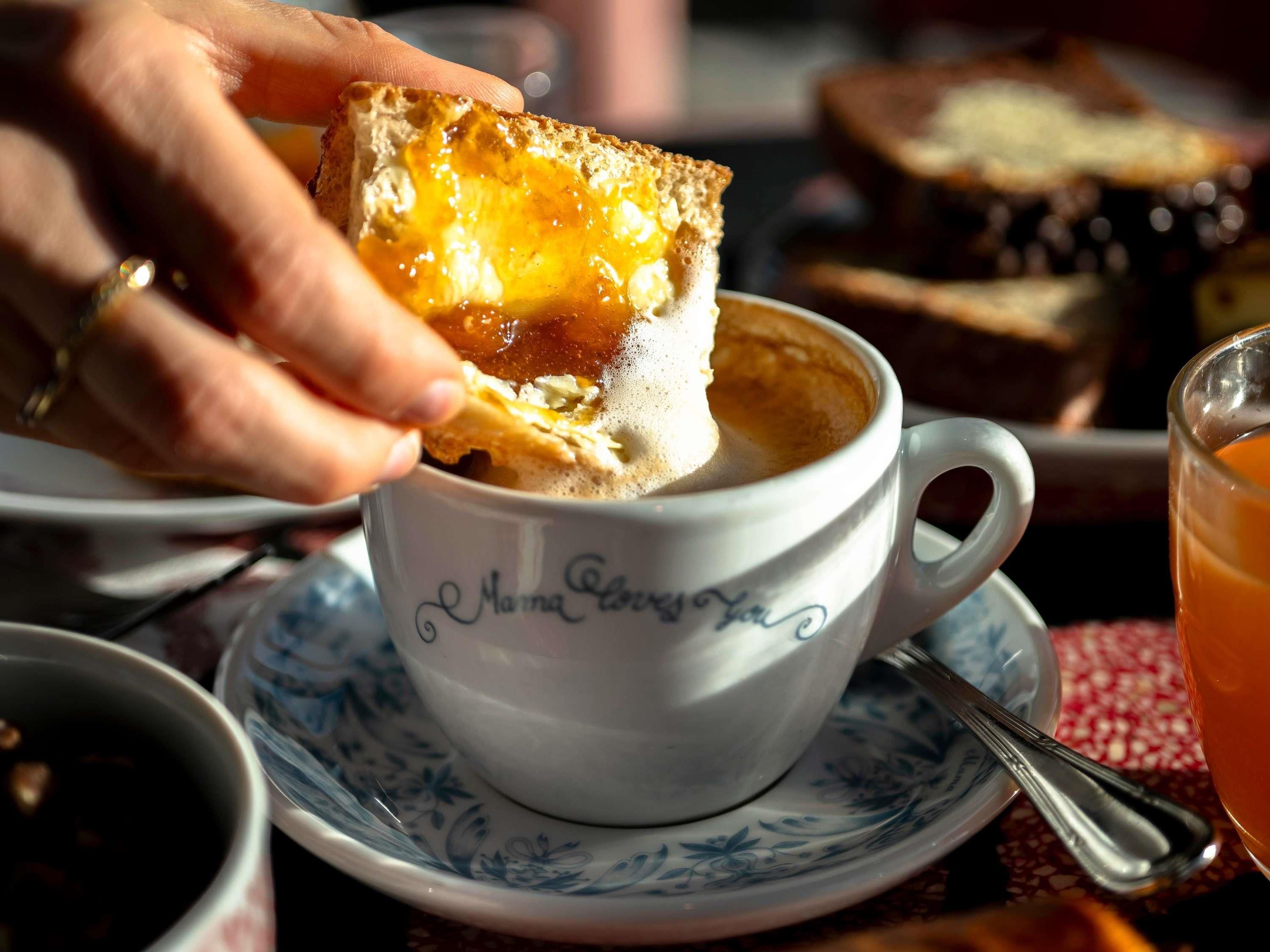 Una taza de café con una tostada, una cuchara y un plato con tostadas al fondo.