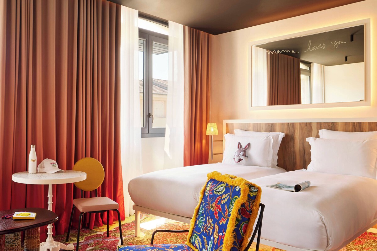 Quarto de hotel com cama, escrivaninha com cadeira, mesa com garrafa e tampa, e espelho com letreiro neon.