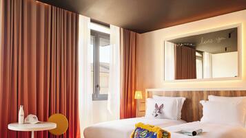 A hotel room with a bed, a desk with a chair, a table with a bottle and cap, and a mirror with a neon sign.