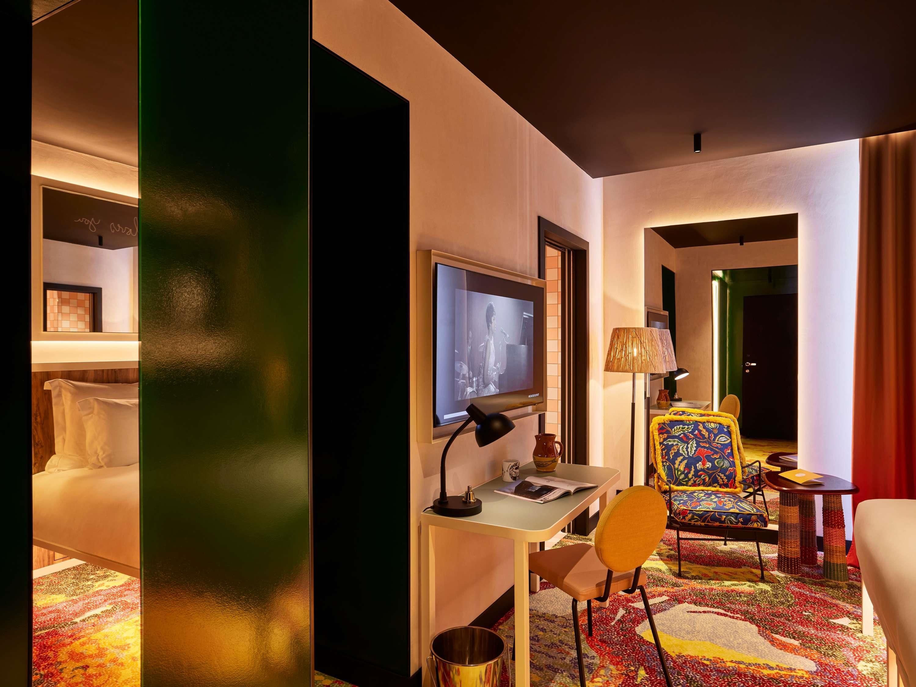 Ein Hotelzimmer mit einem Bett, einem Schreibtisch mit Fernseher, einem Sessel und einer Lampe, sowie einem bunten Teppich.