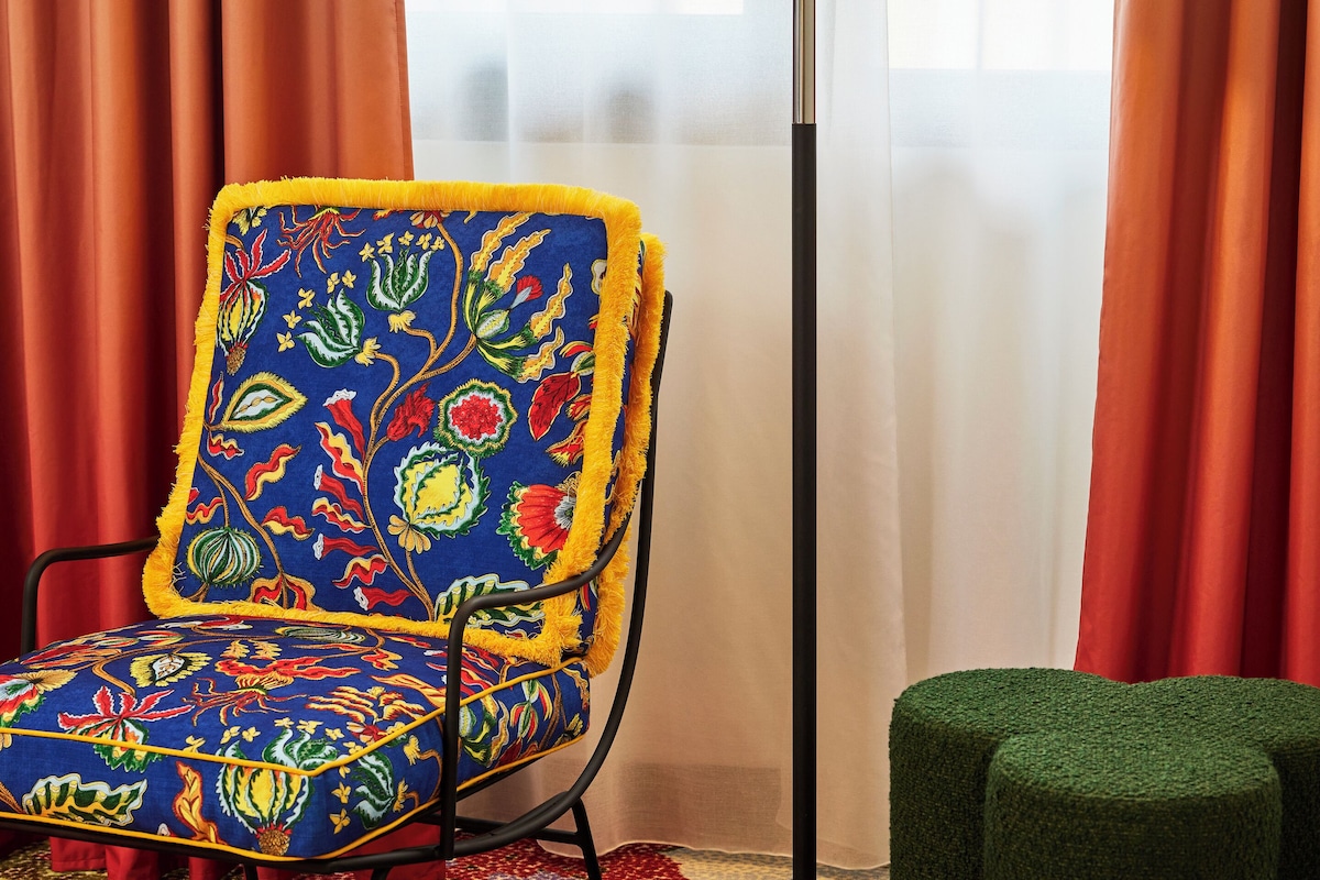 Um quarto com uma poltrona estampada, uma luminária de chão, uma pequena mesa redonda com um livro e uma caneca, e um banquinho verde.