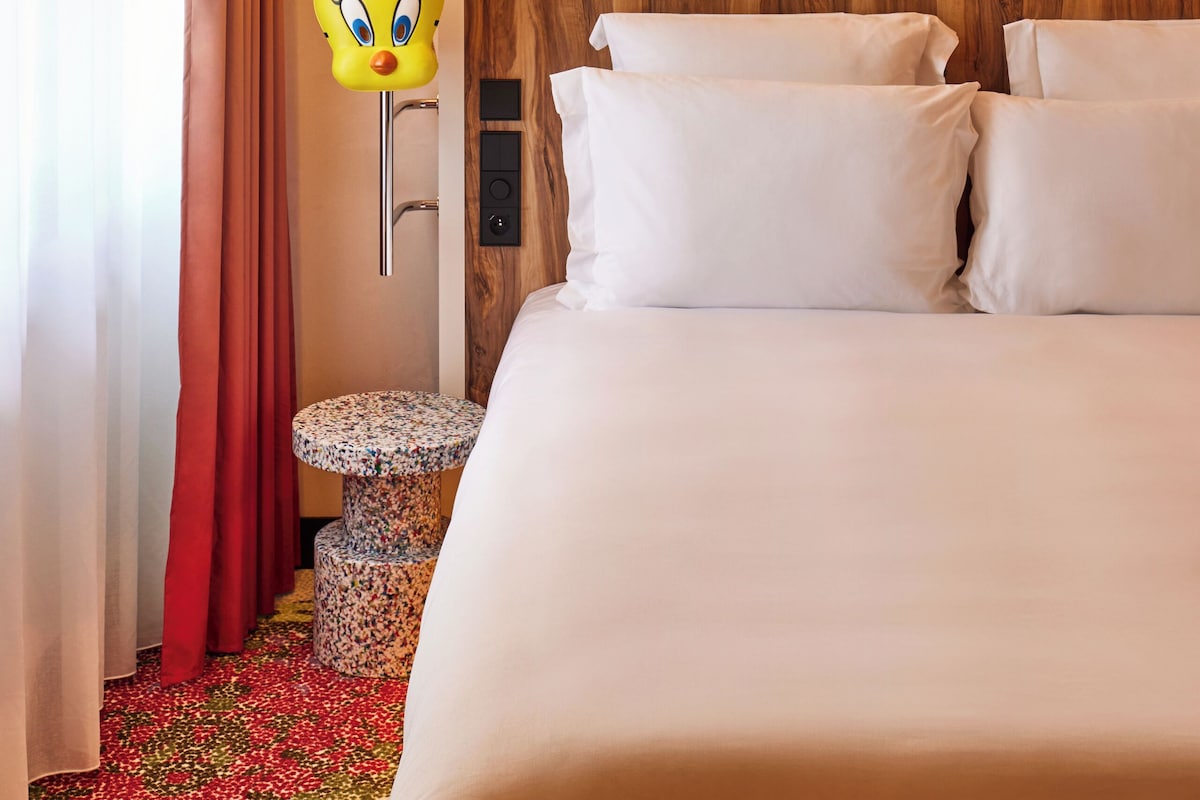 Quarto de hotel com uma cama grande, um espelho, uma mesinha redonda e um adorno decorativo na parede.