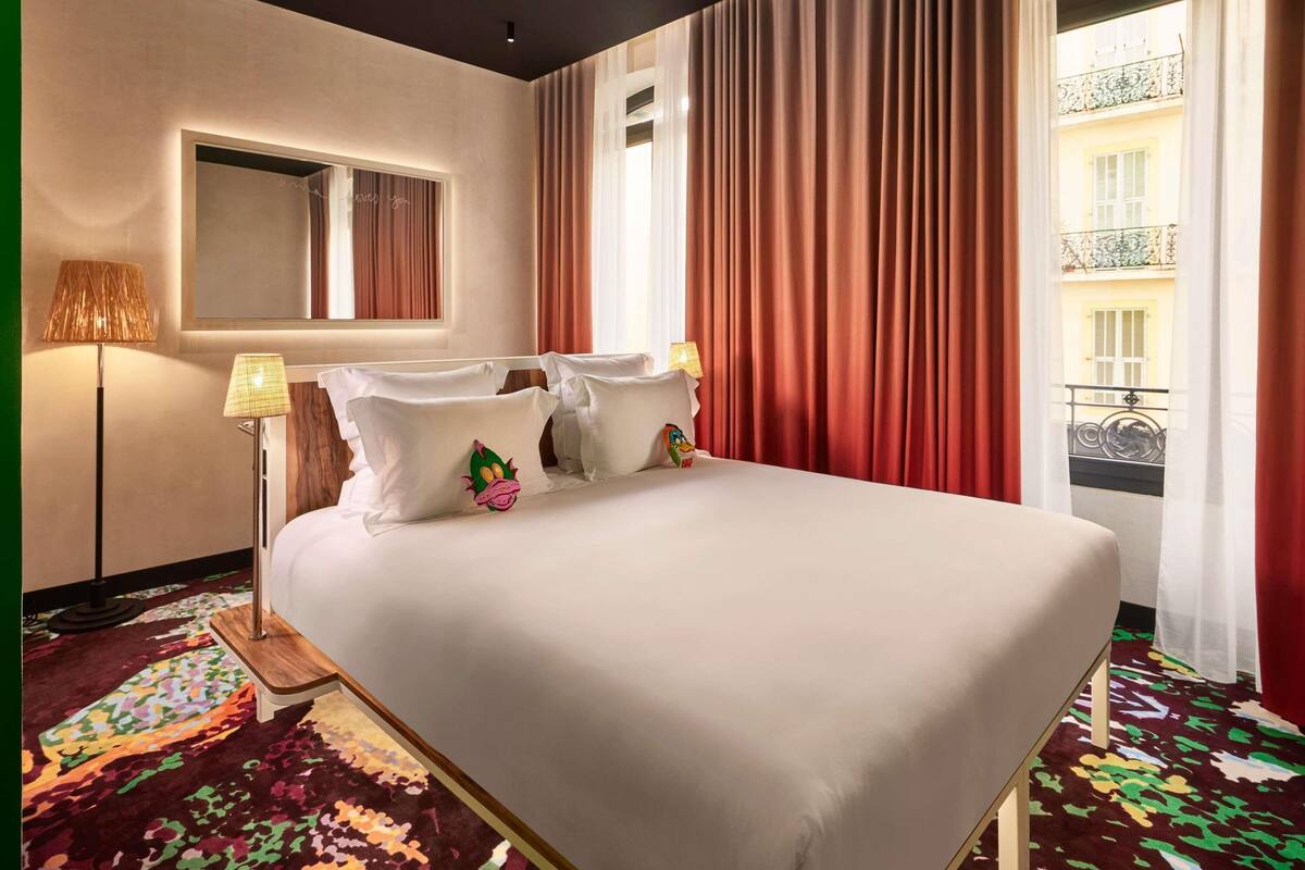 Ein Hotelzimmer mit Bett, zwei Kissen, einer Lampe, einem Spiegel und Blick auf ein Gebäude durch Vorhänge.