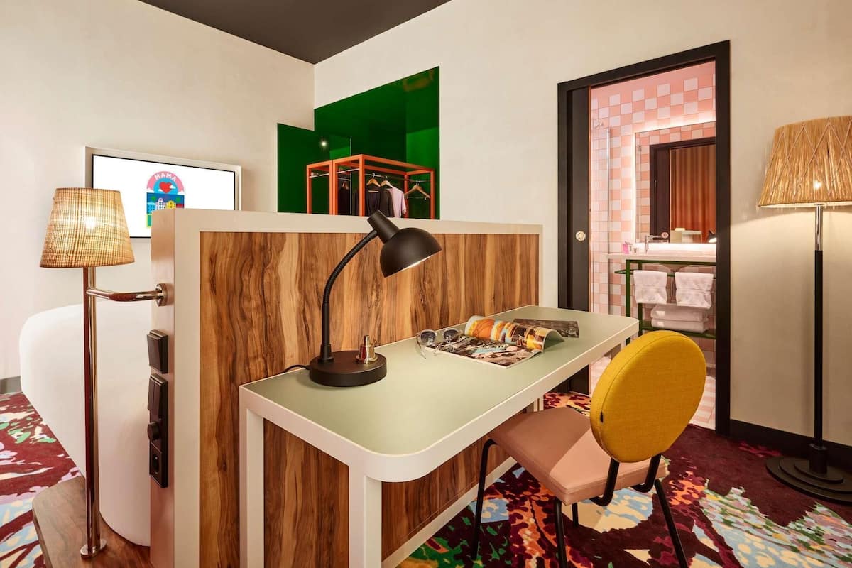 A room with a wooden partition, a desk with a lamp, a chair, a colorful rug, and a bathroom with a tiled wall.