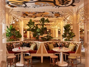 A cozy indoor seating area with round tables and cushioned benches, surrounded by decorative plants and intricate ceiling designs. - Mama Shelter Nice (Nice)