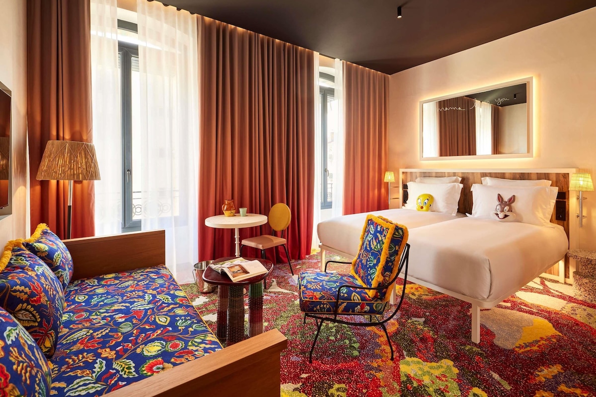 Quarto de hotel com cama, sofá, cadeira, mesa pequena e piso coberto por carpete.