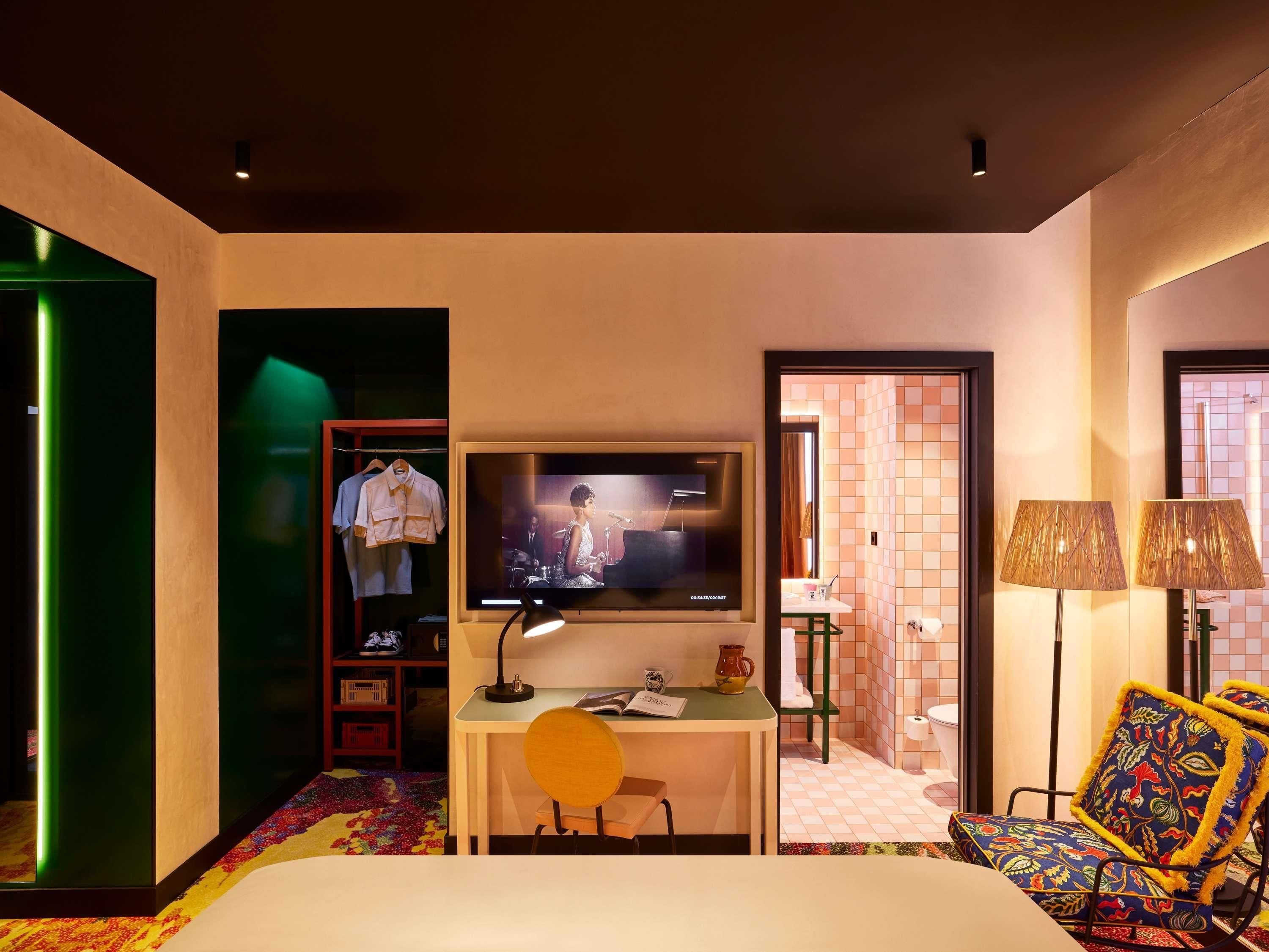 A hotel room with a bed, a desk with a TV, a wardrobe, a chair, and a bathroom visible through an open door.