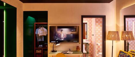 A hotel room with a bed, a desk with a TV, a wardrobe, a chair, and a bathroom visible through an open door.