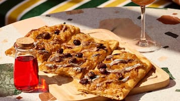 Uma tábua de cortar de madeira com uma fatia de pizza coberta com azeitonas e anchovas, um copo de vinho tinto e uma garrafa pequena de molho de tomate.