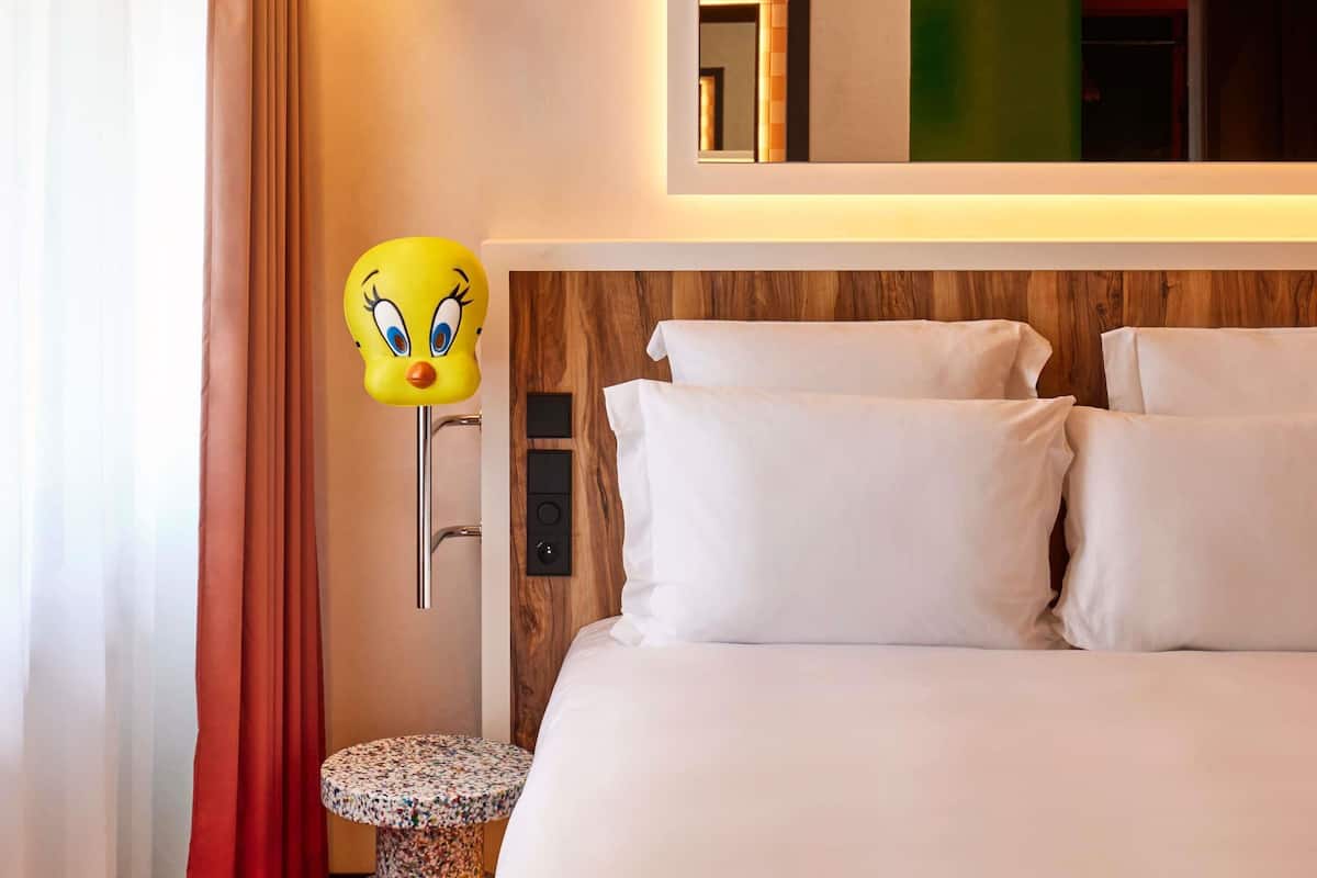 A hotel room with a bed, pillows, a wall-mounted mirror, a small round table, and a decorative headpiece.