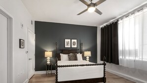 Un dormitorio con pared oscura, cama con dosel de madera, dos lámparas de noche, ventilador de techo y una ventana con cortinas.