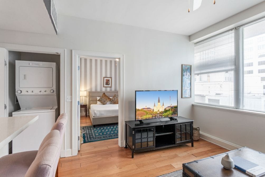 A modern hotel room with a flat-screen TV, a bed with striped curtains, a dining area, and a large window with blinds.