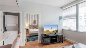 A modern hotel room with a flat-screen TV, a bed with striped curtains, a dining area, and a large window with blinds.