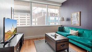 Een moderne woonkamer met een groot raam, een groene bank, een houten salontafel en een televisie op een zwart tv-meubel.