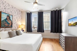 Una habitación de hotel moderna con una cama grande, un ventilador de techo, vistas a la ciudad a través de la ventana y un televisor de pantalla plana en la pared.