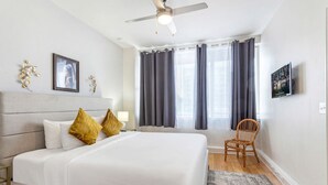 Une chambre d’hôtel avec un grand lit, un ventilateur de plafond, une fenêtre avec des rideaux, une chaise et un téléviseur fixé au mur.