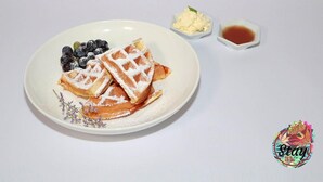 Une assiette avec des gaufres, des myrtilles et du sucre glace, accompagnée de deux petits bols contenant du beurre et du sirop.
