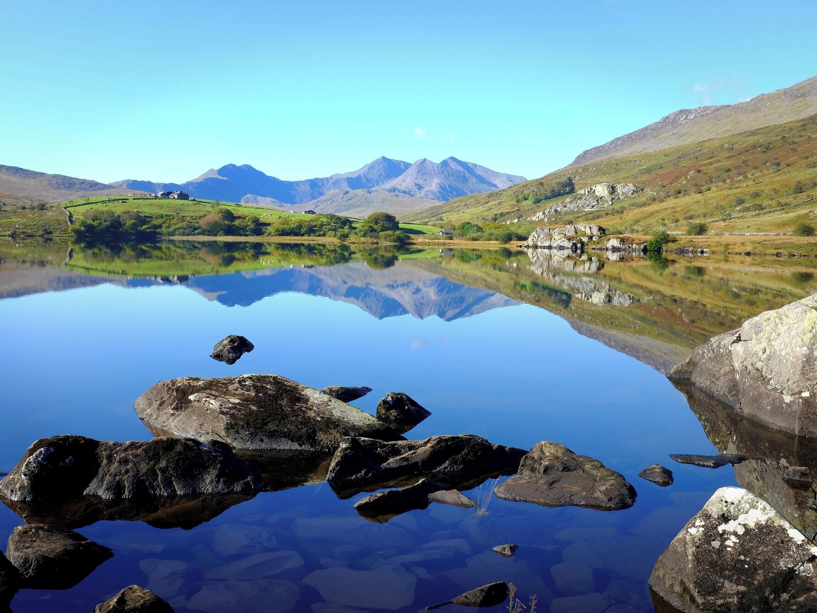 A tranquil lake with clear reflections of surrounding mountains and a small house on the shore.