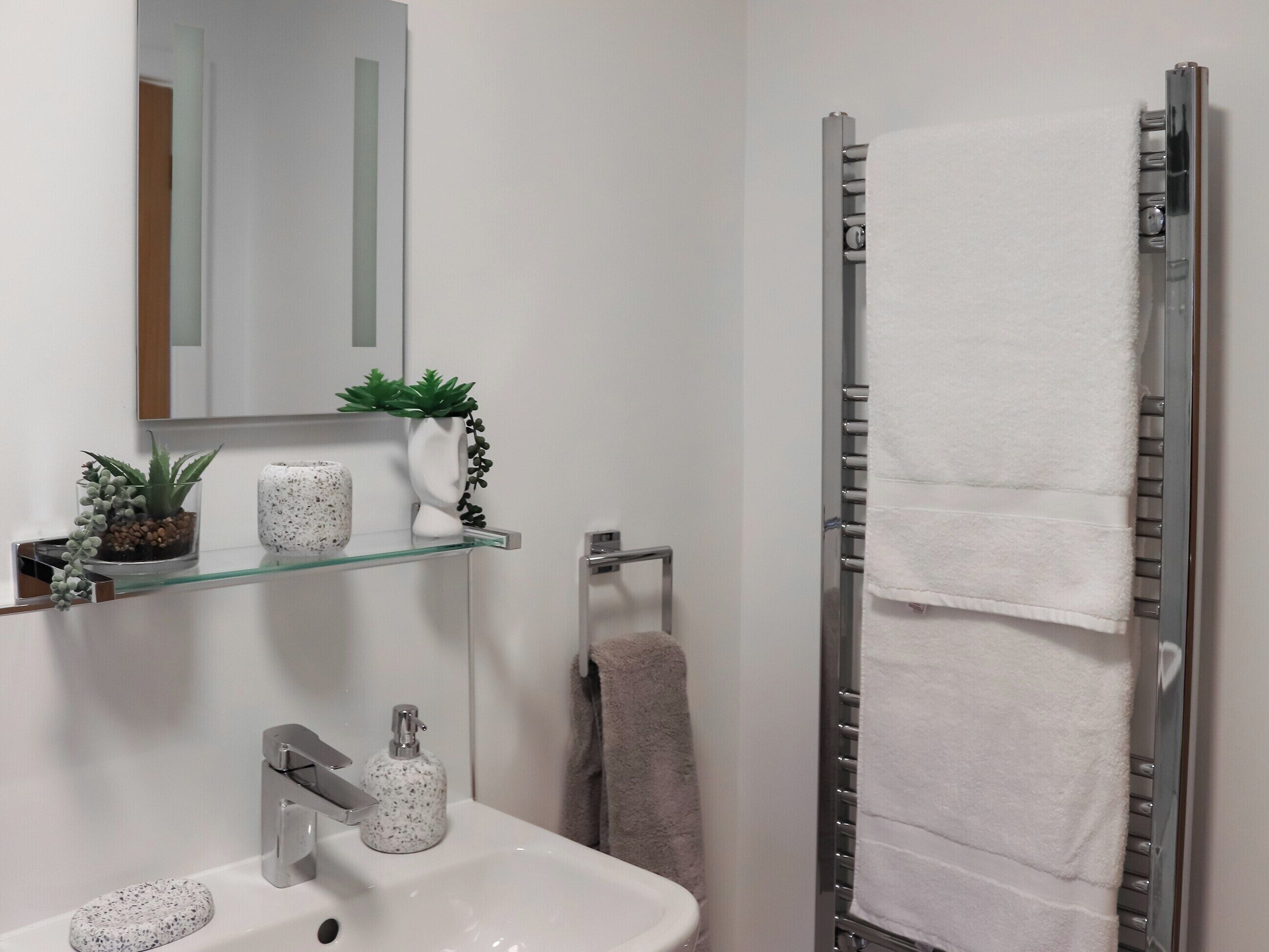 Une salle de bain avec un lavabo blanc, un miroir, une étagère en verre avec des plantes, un porte-serviettes avec une serviette blanche et une douche avec une porte en verre.
