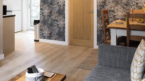 Un salon avec un canapé, une table basse sur laquelle sont posées des télécommandes, un coin repas avec une table et des chaises, une horloge au mur et une porte donnant sur une autre pièce.
