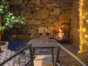 Un muro de piedra, una mesa con dos copas de vino y una pequeña fogata.