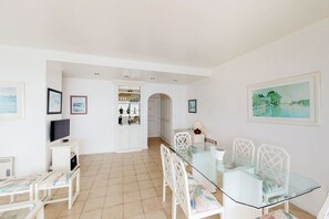 A spacious room with a glass dining table, white chairs, a TV, and a kitchen area in the background.