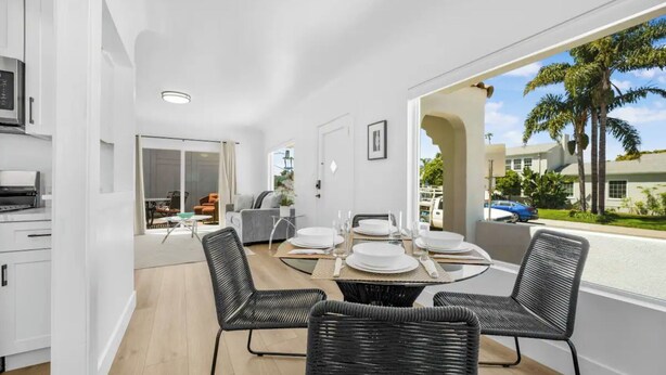 Ein modernes Esszimmer mit einem Glastisch und schwarzen Korbstühlen, eine Küche mit weißen Schränken sowie Blick auf Palmen und eine Straße.