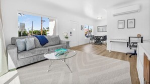 Ein modernes Wohnzimmer mit einem grauen Sofa, einem Glastisch und einem Essbereich mit weißem Tisch und Stühlen.