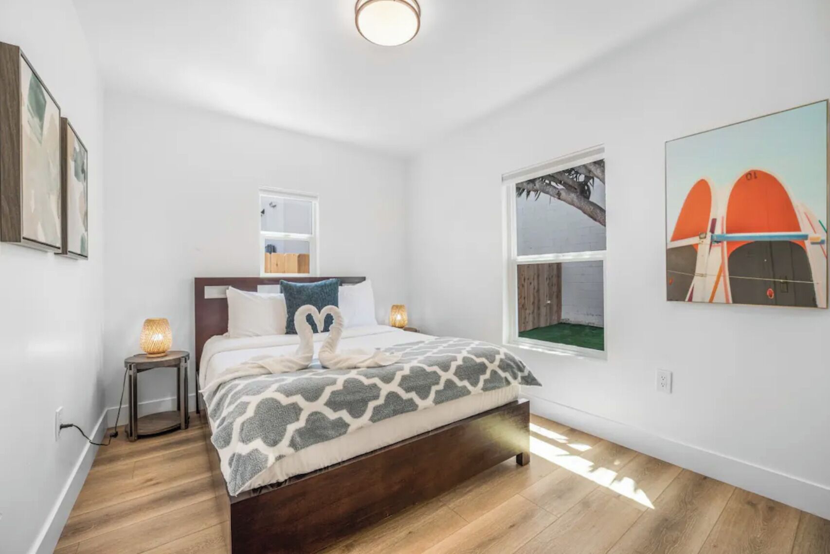 A modern bedroom with a wooden bed, a small bedside table, a nightstand, a window with a view of a tree, and a painting on the wall.
