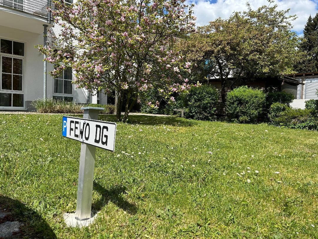 Um sinal de estacionamento com a inscrição 'FEWO DG' numa área ajardinada, com um edifício ao fundo.