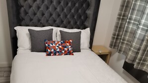 Una cama bien tendida con un cabecero capitonado, dos almohadas, una colcha y una mesita de noche con un control remoto.