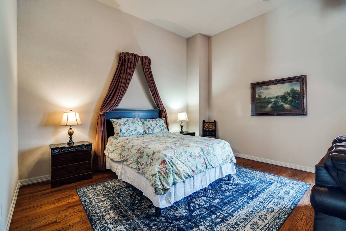 A bedroom with a bed, two bedside tables, a nightstand, a blue patterned rug, a wooden floor, a painting on the wall, and a dark-colored chair.