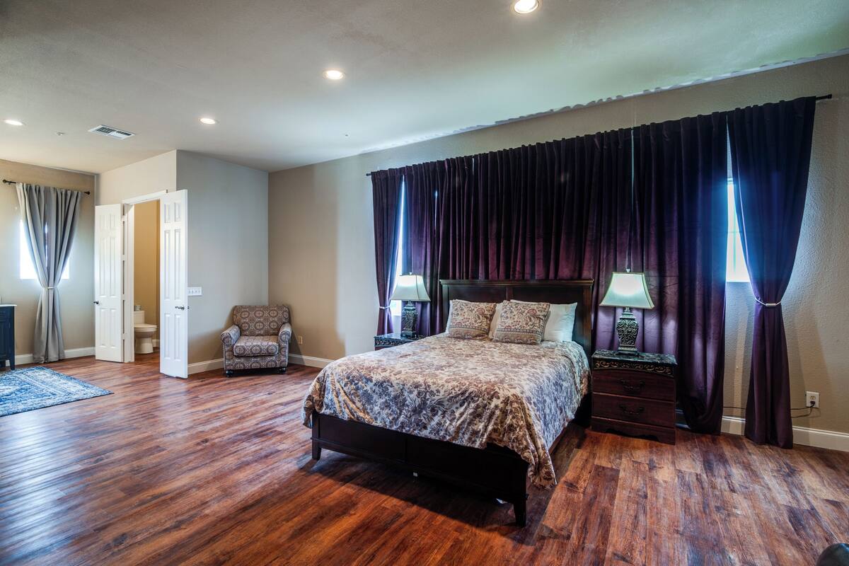 A bedroom with a large bed, a chair, a nightstand, and a window with curtains.