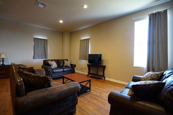 A living room with a brown leather sofa, a wooden coffee table, a television on a stand, and a window with curtains. - The Castle at Rockwall (Rockwall)