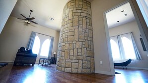 A spacious living room with a stone fireplace, a ceiling fan, and large windows.