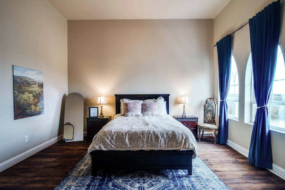 A bedroom with a large bed, two bedside lamps, a chair, a mirror, and a window with curtains.