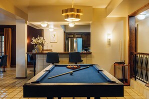 Une table de billard avec des boules et des queues, un coin bar avec des chaises, et une cuisine équipée d’un réfrigérateur.