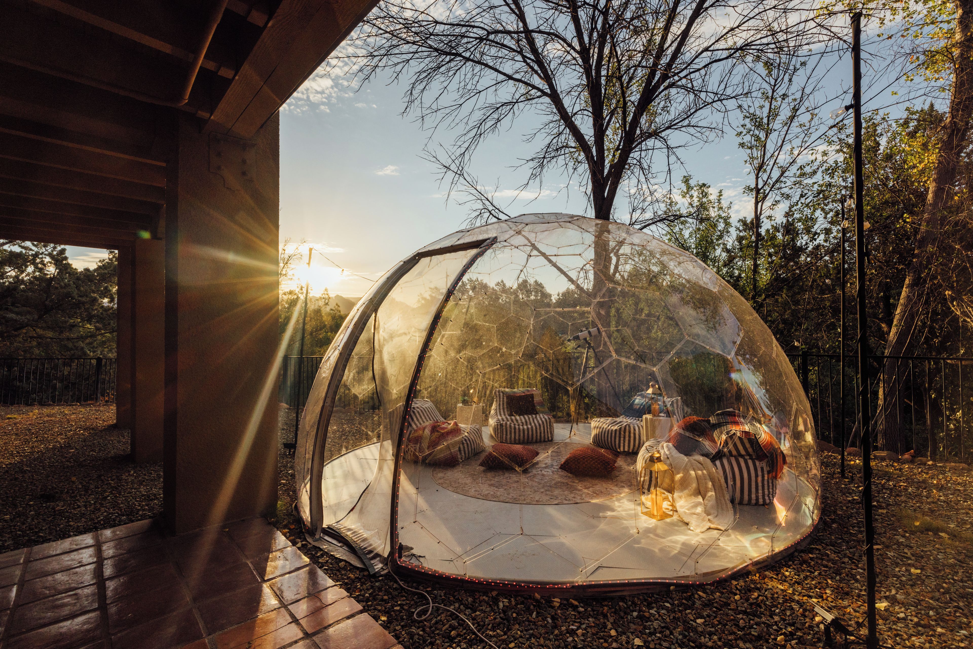 A transparent dome structure with seating inside, set in an outdoor environment with trees and a wooden building in the background.