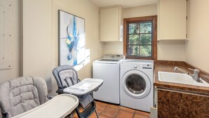 A laundry room with a washing machine, dryer, and a sink. There is a chair with a tray and a window with a view of trees.