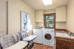 A laundry room with a washing machine, dryer, and a sink. There is a chair with a tray and a window with a view of trees.