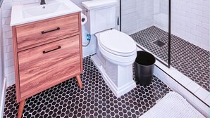 Moderni kylpyhuone, jossa on valkoinen pesuallas, puinen allaskaappi, wc ja lasiseinällä erotettu suihkutila.